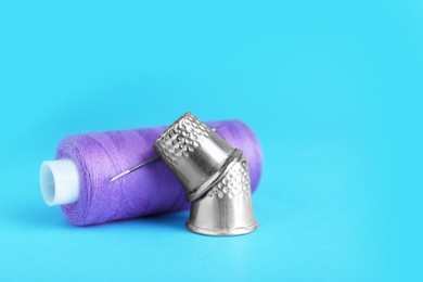 Thimbles and spool of purple sewing thread with needle on light blue background, closeup. Space for text