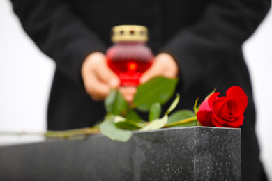 Woman with candle near black granite tombstone outdoors, focus on red rose. Funeral ceremony