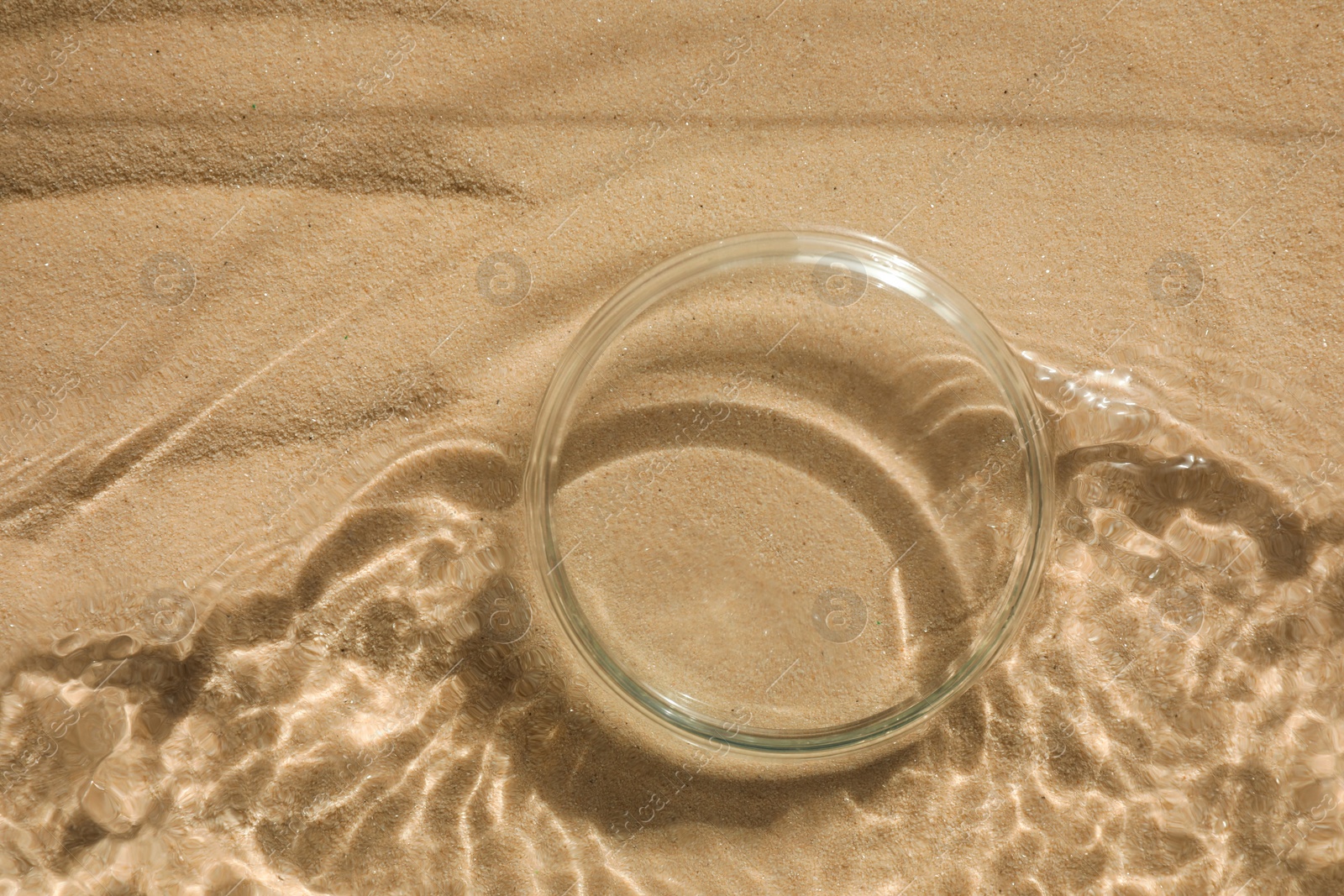 Photo of Stylish presentation for product. Glass podium in water on sand, top view