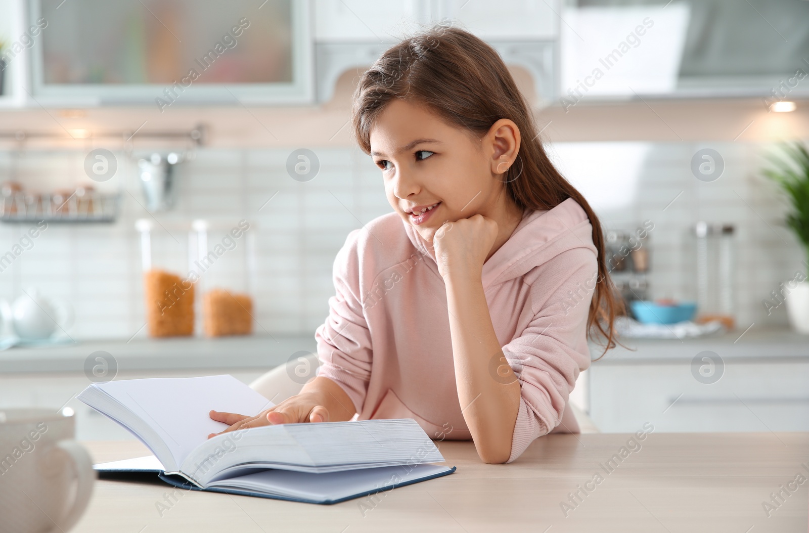 Photo of Cute little girl reading book at table in kitchen