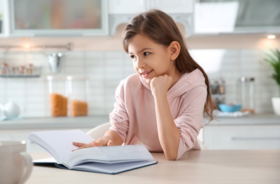 Cute little girl reading book at table in kitchen