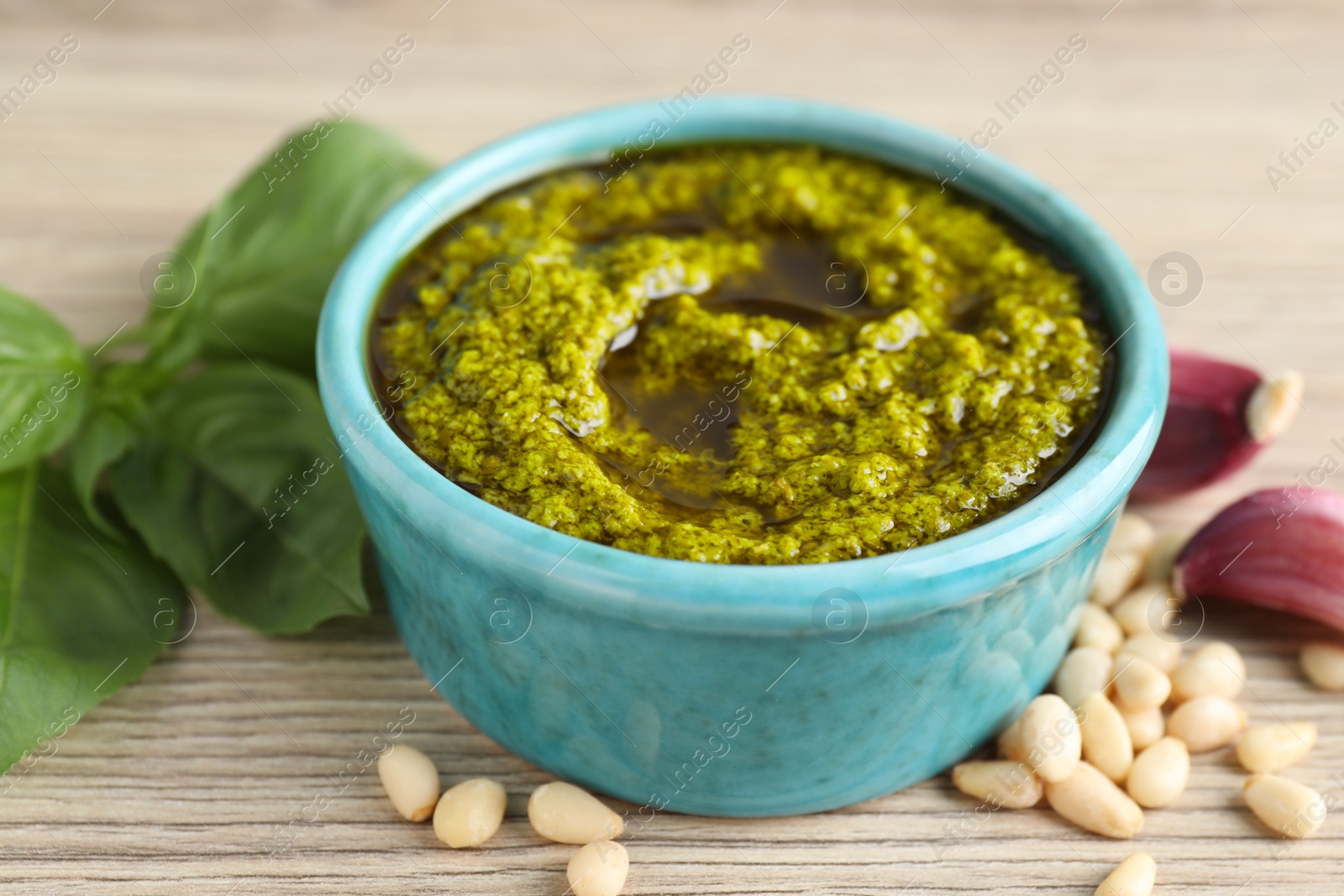 Photo of Tasty pesto sauce in bowl, pine nuts, garlic and basil on wooden table, closeup