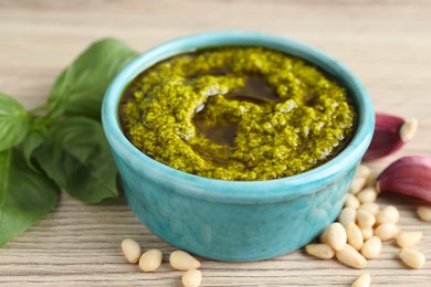 Photo of Tasty pesto sauce in bowl, pine nuts, garlic and basil on wooden table, closeup