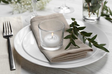 Stylish setting with cutlery, burning candle and eucalyptus leaves on white wooden table, closeup