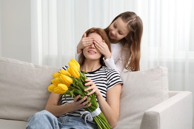 Photo of Daughter covering mother's eyes with her palms and congratulating with bouquet of yellow tulips at home