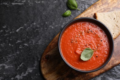 Delicious tomato cream soup in bowl and basil leaves on dark textured table, flat lay. Space for text