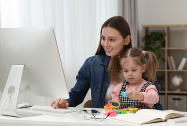 Photo of Woman working remotely at home. Mother using computer while her daughter playing at desk