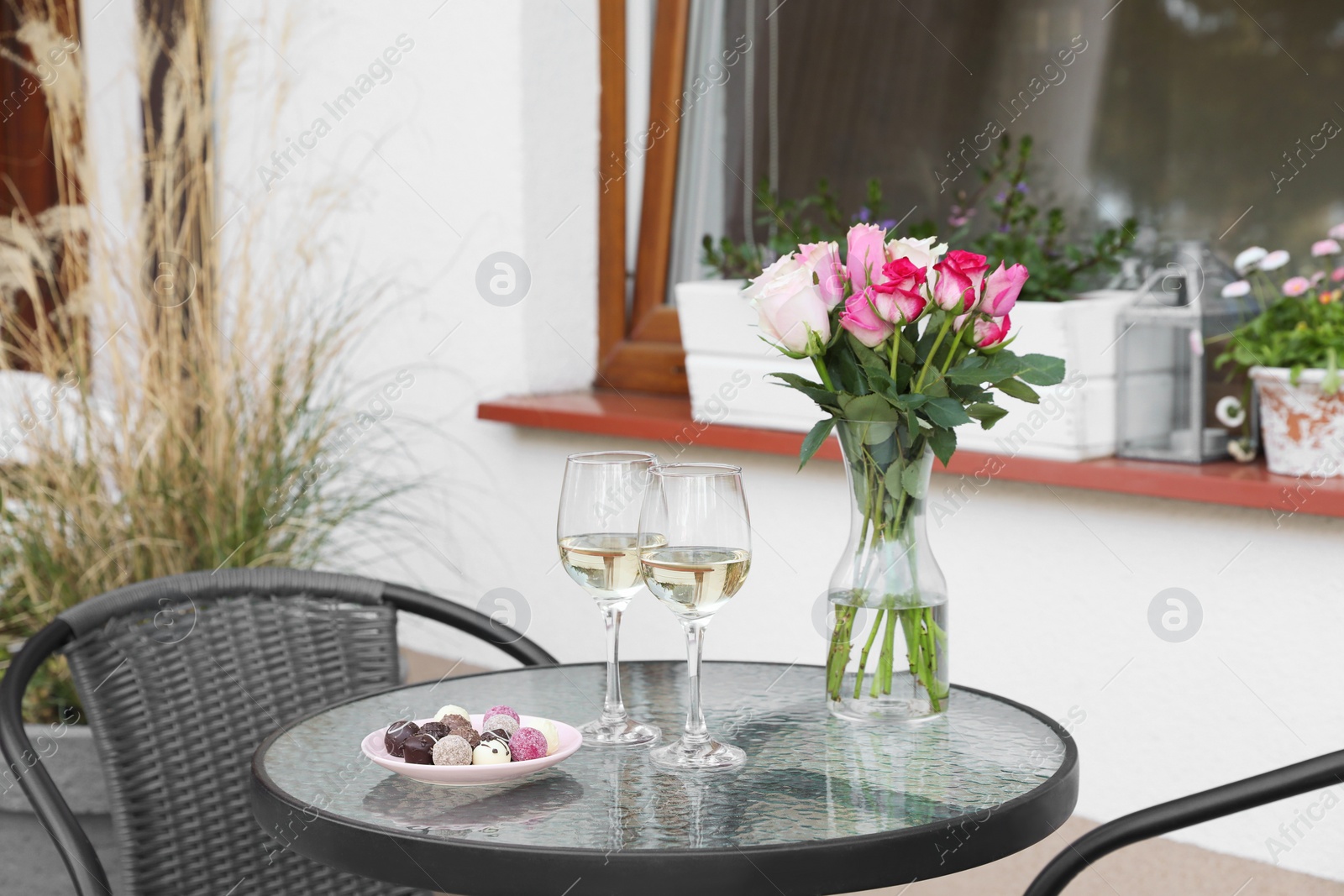 Photo of Vase with roses, glasses of wine and candies on glass table near house on outdoor terrace