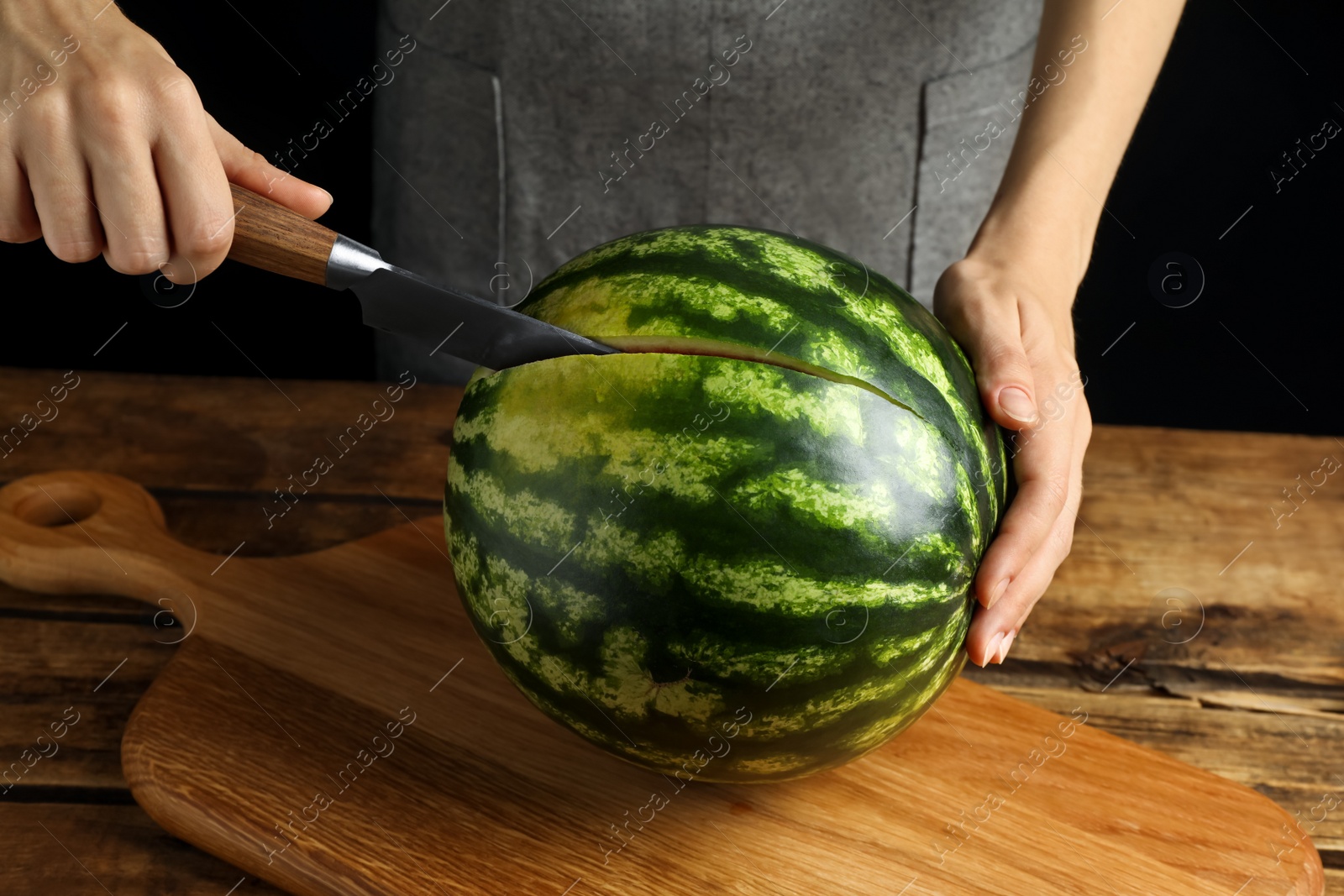 Photo of Woman cutting delicious watermelon at wooden table against black background, closeup