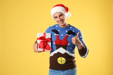 Photo of Happy man in Christmas sweater and Santa hat holding gift box on yellow background