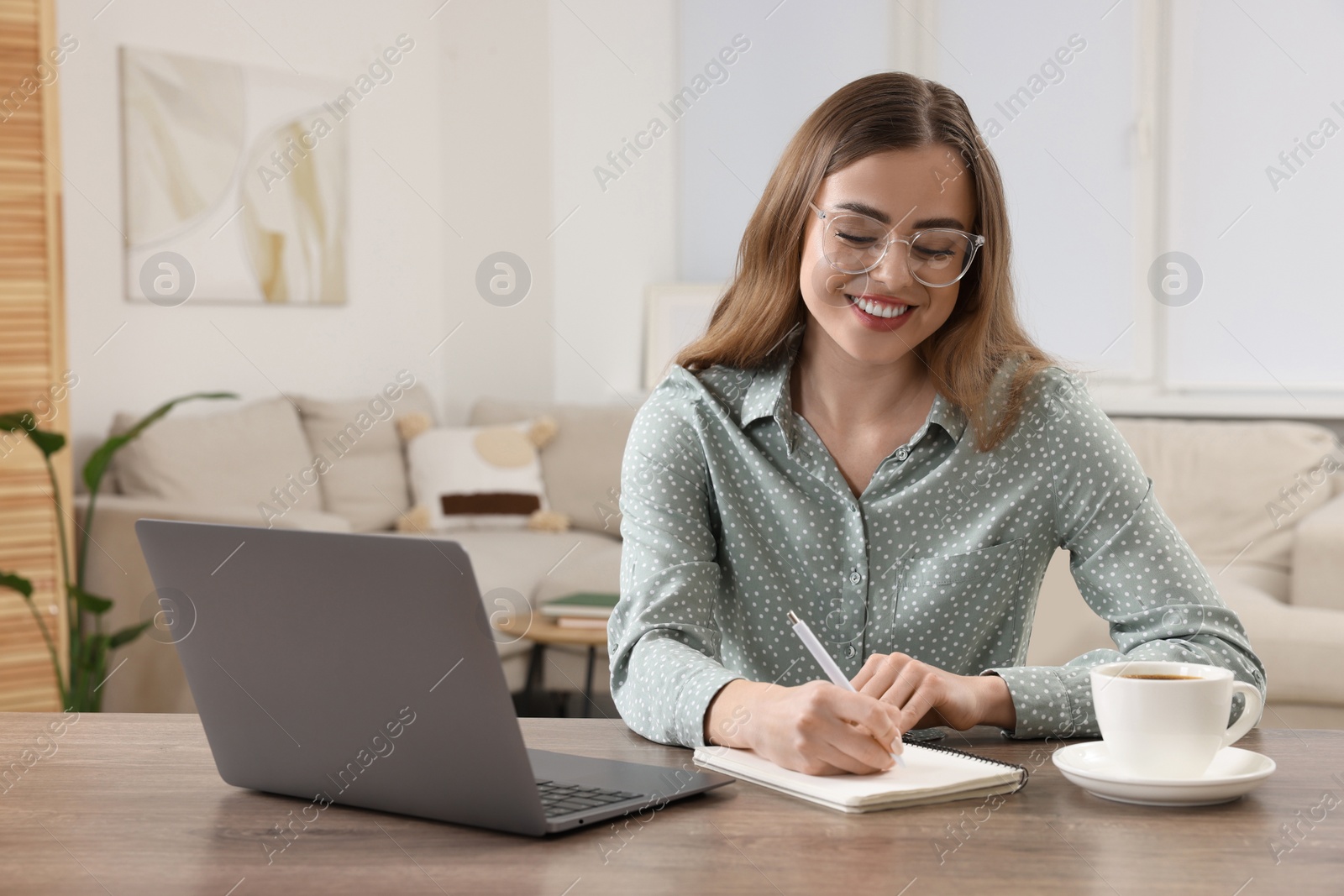 Photo of Happy woman writing something in notebook near laptop at wooden table in room