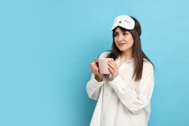 Woman in pyjama and sleep mask holding cup of drink on light blue background, space for text