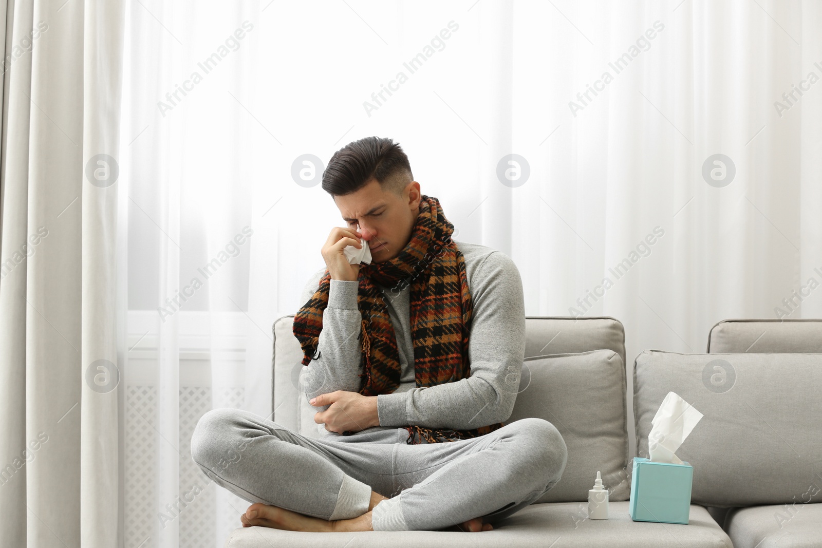 Photo of Ill man sitting near nasal spray and box of paper tissues on sofa at home