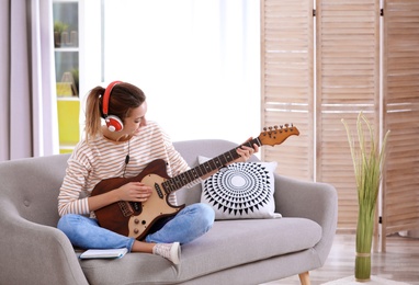 Young woman with headphones playing electric guitar in living room. Space for text