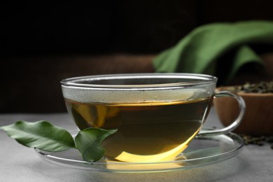Photo of Fresh green tea in glass cup with saucer and leaves on grey table, closeup