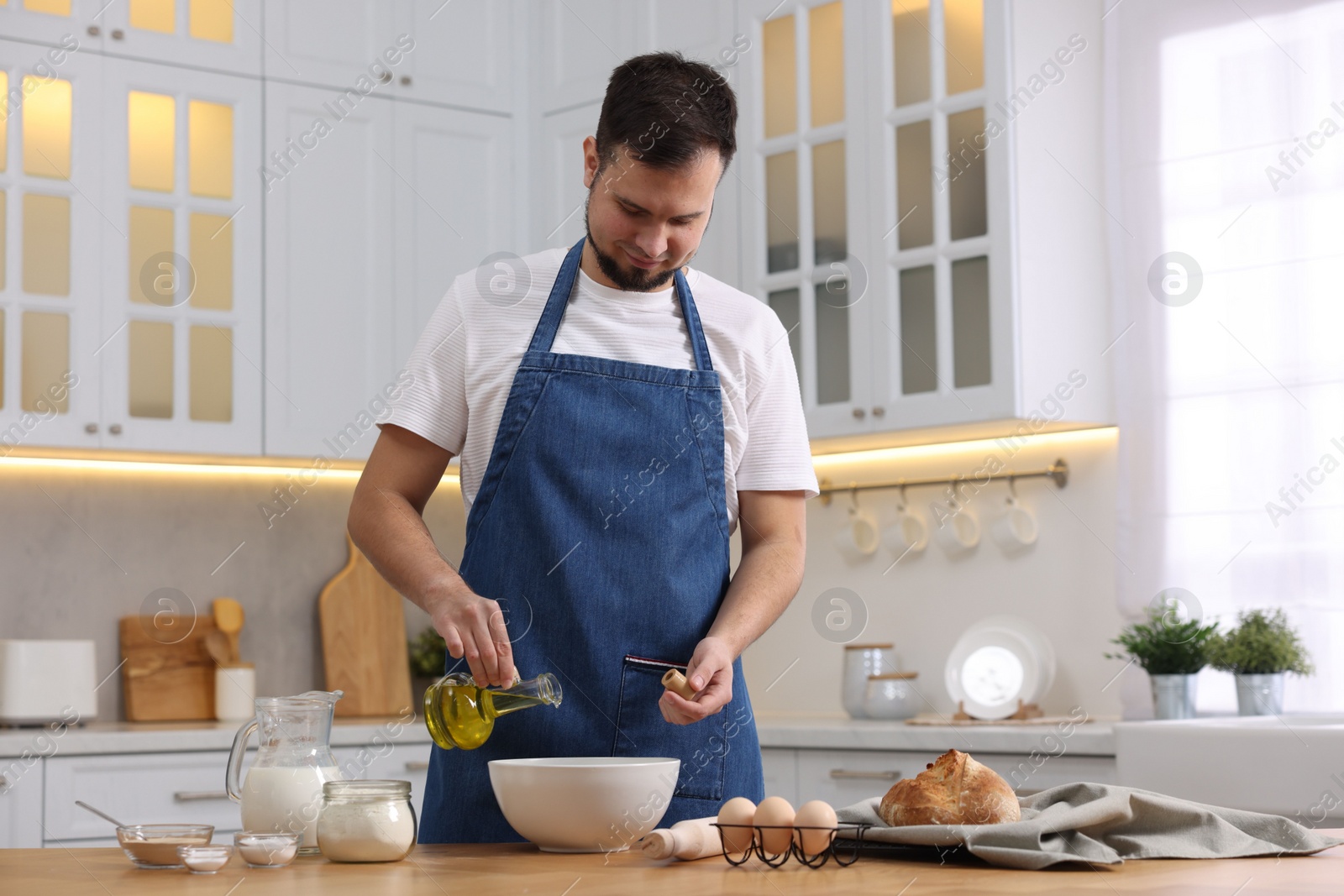 Photo of Making bread. Man pouring oil into bowl at wooden table in kitchen