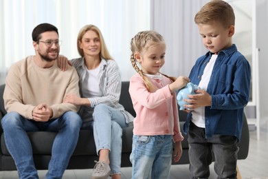 Photo of Family budget. Children putting coin into piggy bank while their parents watching at them indoors, selective focus