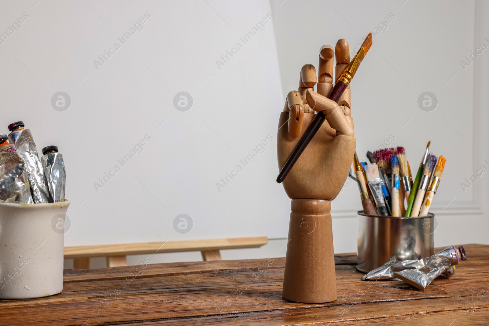 Photo of Easel with blank canvas, hand model and different art supplies on wooden table near white wall