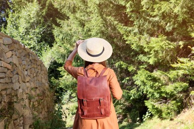 Photo of Woman with backpack and hat walking along stone wall in forest, back view