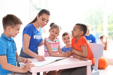 Young volunteer reading book with children at table indoors