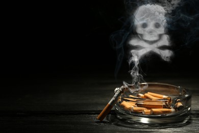 Image of No Smoking. Skull and crossbones symbol of smoke over ashtray with stubs and smoldering cigarette on dark wooden table against black background. Space for text