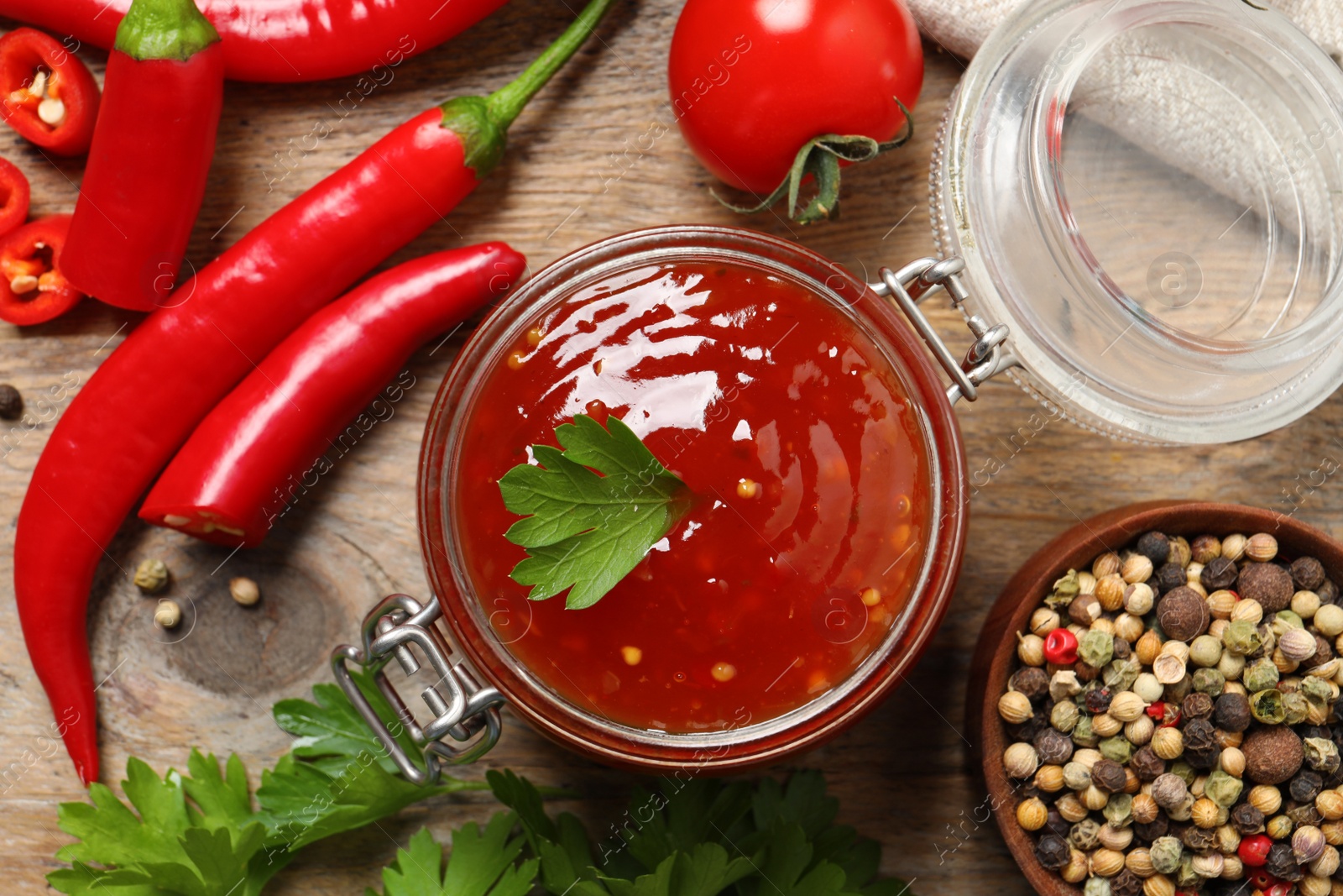 Photo of Spicy chili sauce in glass jar and ingredients on wooden table, flat lay