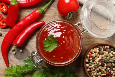 Photo of Spicy chili sauce in glass jar and ingredients on wooden table, flat lay