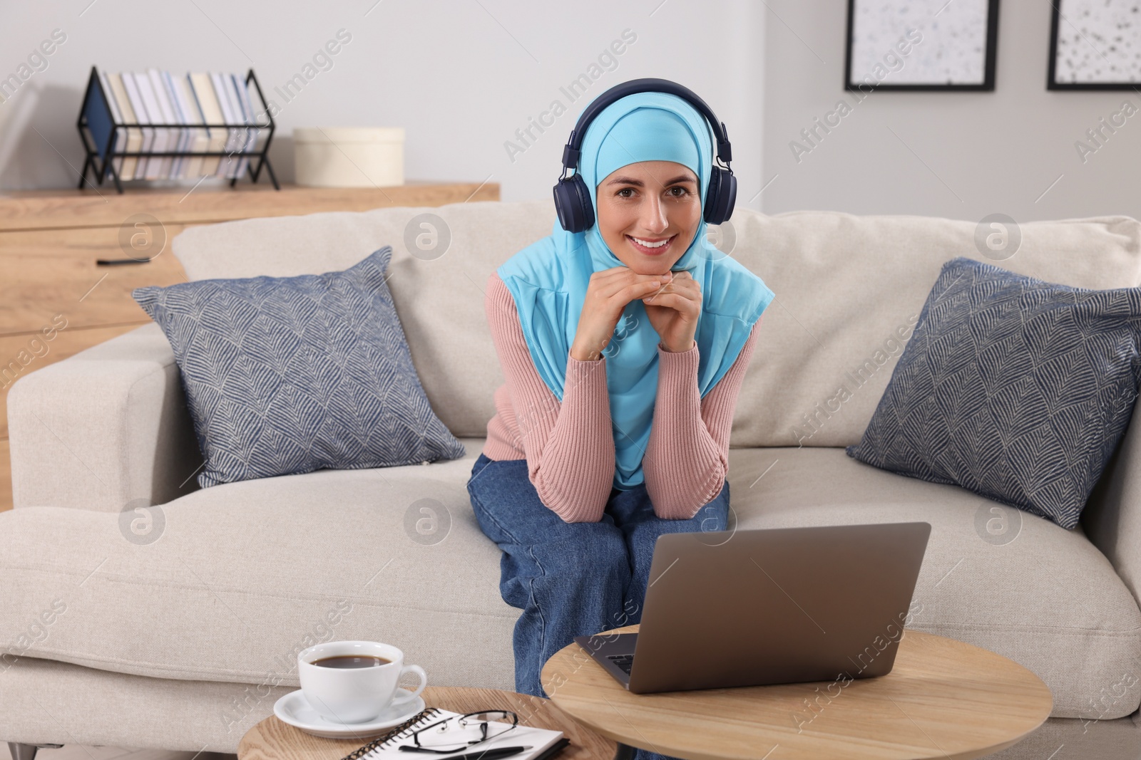 Photo of Muslim woman in headphones using laptop at wooden table in room. Space for text