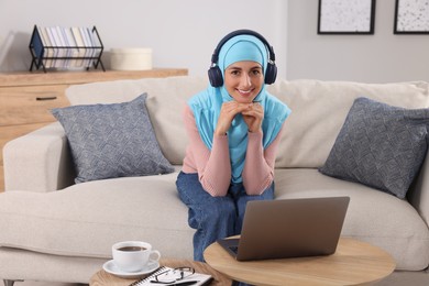 Muslim woman in headphones using laptop at wooden table in room. Space for text