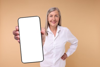 Happy mature woman showing mobile phone with blank screen on beige background. Mockup for design