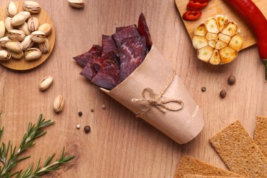 Paper bag with pieces of delicious beef jerky and different spices on wooden table, flat lay