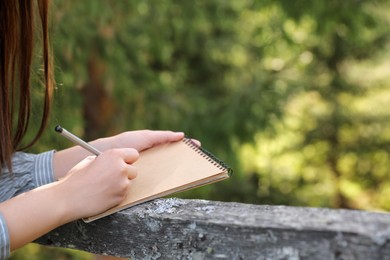 Photo of Young woman drawing with pencil in notepad near wooden fence, closeup