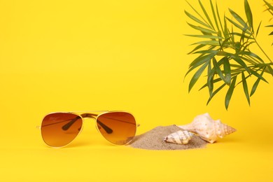Photo of Stylish sunglasses, seashells, sand and palm leaves on yellow background