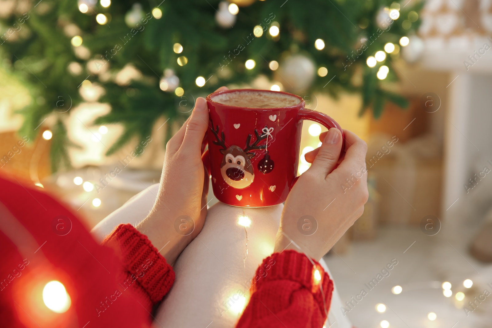 Photo of Woman holding festive cup with coffee near Christmas tree indoors, closeup