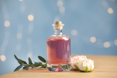 Bottle of essential oil, roses and green branch on wooden table against blurred lights