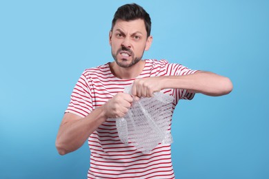 Angry man popping bubble wrap on light blue background. Stress relief