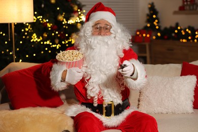 Photo of Merry Christmas. Santa Claus with popcorn bucket changing TV channels on sofa at home