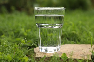 Photo of Glass of fresh water on stone in green grass outdoors, closeup