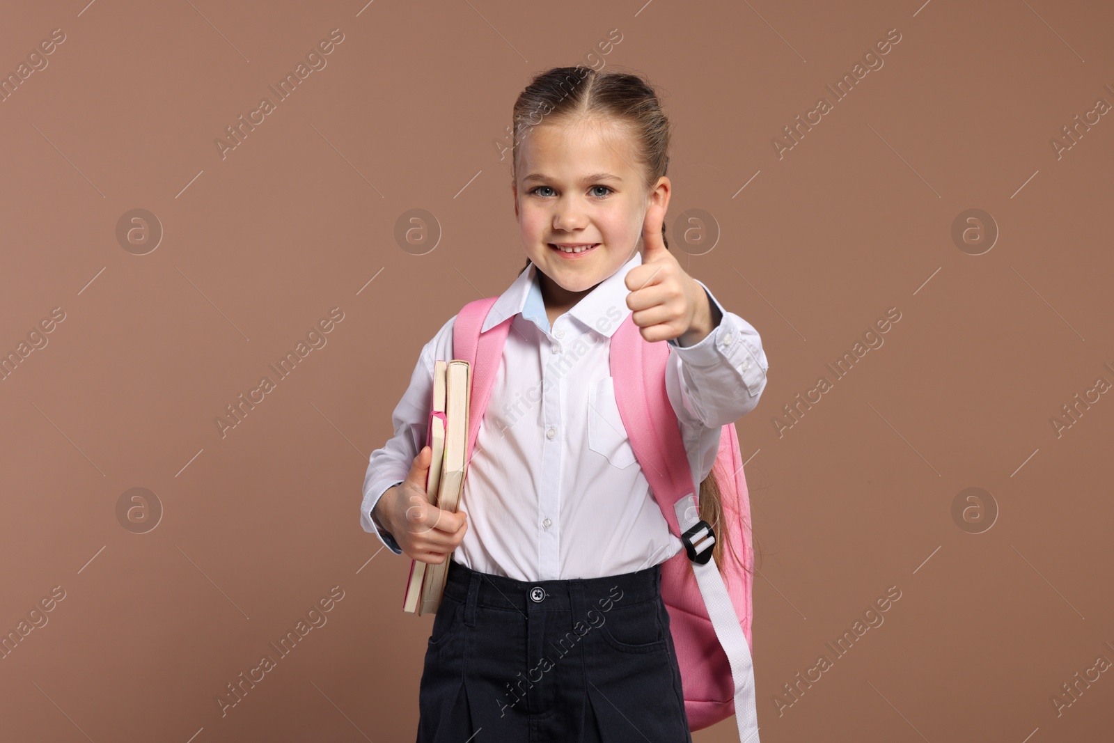 Photo of Happy schoolgirl with backpack and books showing thumb up gesture on brown background