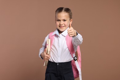 Happy schoolgirl with backpack and books showing thumb up gesture on brown background