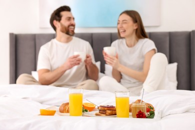 Photo of Tray with tasty breakfast on bed. Happy couple laughing in bedroom, selective focus