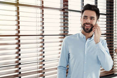 Handsome man talking on mobile phone at window indoors. Space for text