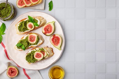 Tasty bruschettas with cream cheese, pesto sauce, figs and fresh basil on white tiled table, flat lay. Space for text