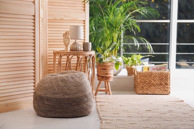 Photo of Stylish knitted pouf, wicker furniture and beautiful houseplants in room. Interior design