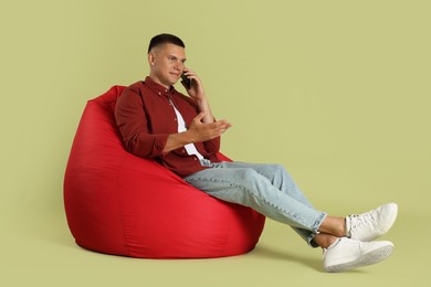 Photo of Handsome man talking by smartphone on red bean bag chair against green background