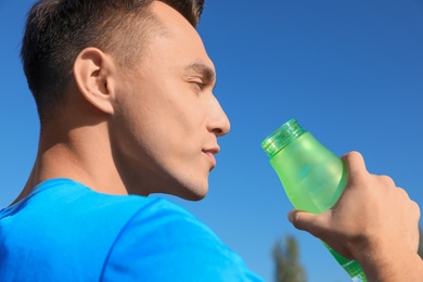 Young sporty man drinking water from bottle against blue sky on sunny day