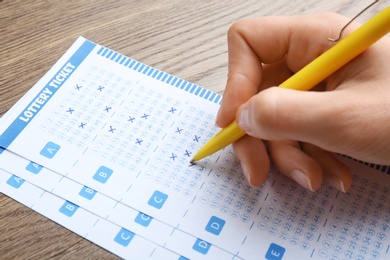 Photo of Woman filling out lottery tickets with pen on wooden table, closeup. Space for text