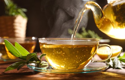Photo of Pouring aromatic herbal tea into cup and fresh sage on wooden table, closeup
