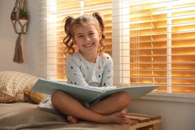 Photo of Cute little girl reading book near window at home