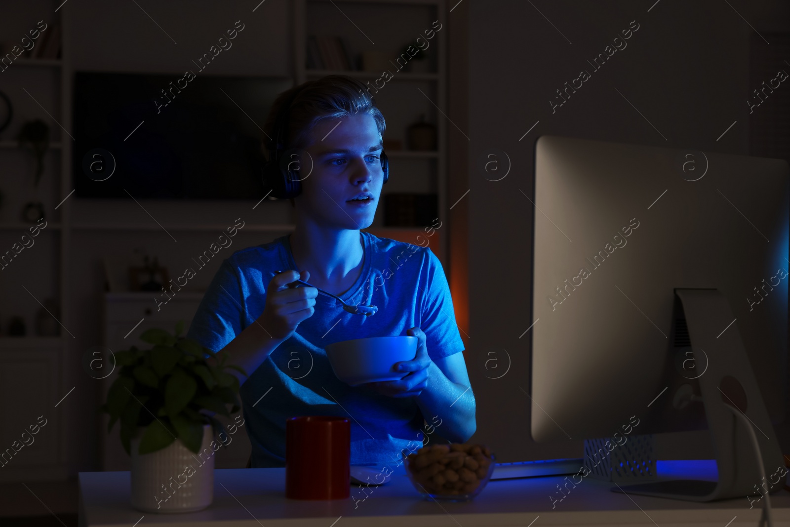 Photo of Teenage boy eating porridge while using computer in room at night. Internet addiction